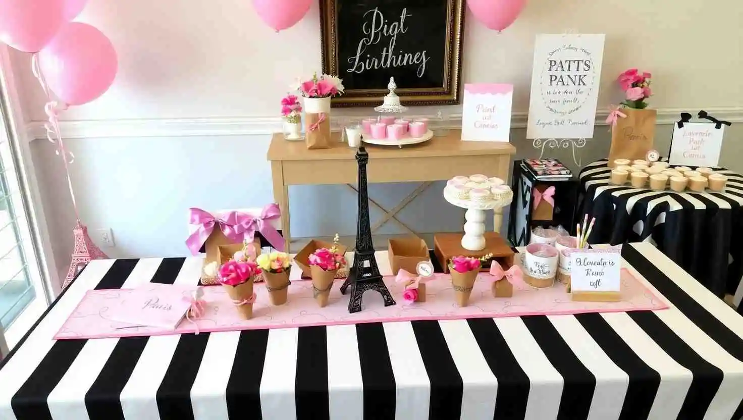 Paris-themed birthday dessert table featuring a black-and-white striped tablecloth, miniature Eiffel Tower centerpiece, floral cones, pink cupcakes, and signs with Paris-inspired designs. Pink balloons and a decorative Eiffel Tower accent complete the setup.