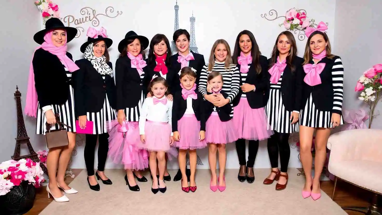 Group photo at a Paris-themed party featuring women and children dressed in coordinated outfits with black and white stripes, pink tutus, bows, and scarves, standing in front of Eiffel Tower decor and floral arrangements.