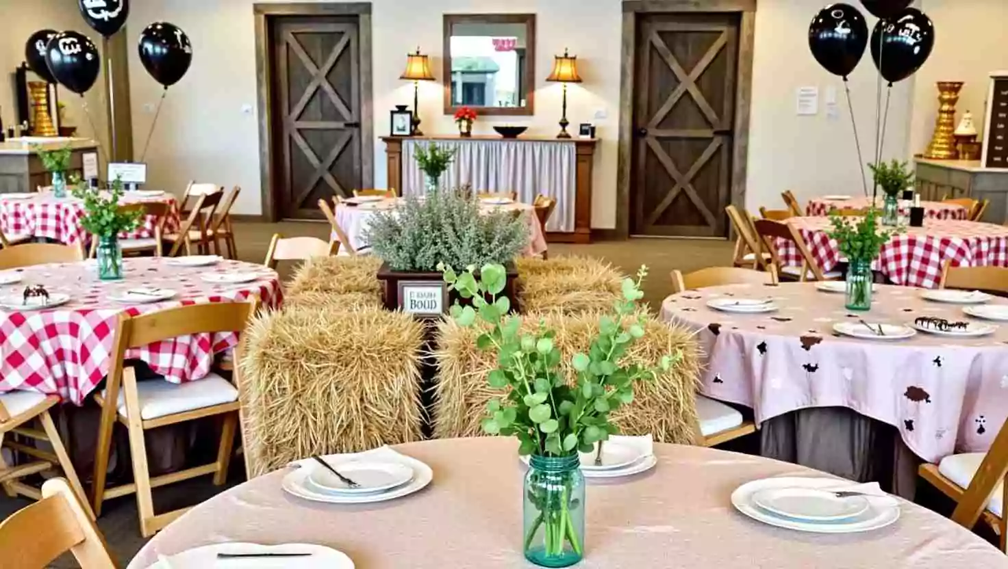 An indoor farm-themed birthday party setup with round tables covered in gingham and farm-printed tablecloths. The decor features hay bales, green plants in mason jars, and black farm-themed balloons. The barn-style doors and cozy lighting add a charming, rustic atmosphere.