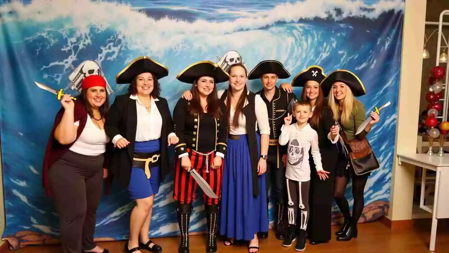 A group of adults and a child posing in front of a pirate-themed backdrop, wearing pirate costumes with hats and props. A fun photo moment at a pirate-themed event.